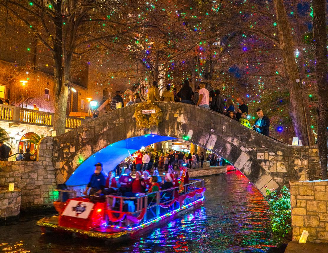 A River Walk boat decorated with lights passing beneath a stone bridge at night